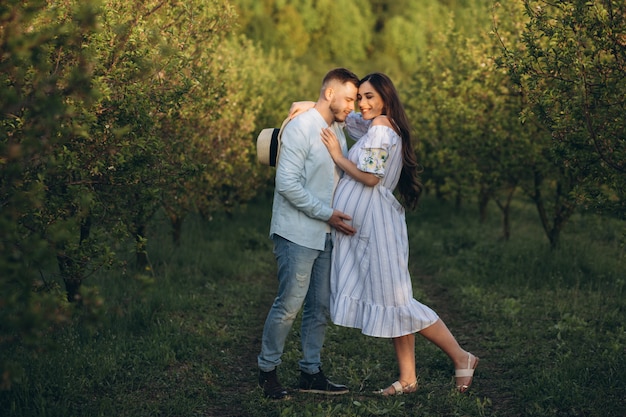 La donna incinta felice alla moda ed alla moda e suo marito hanno vestito un tono bianco e blu pastello nel giardino sul tramonto