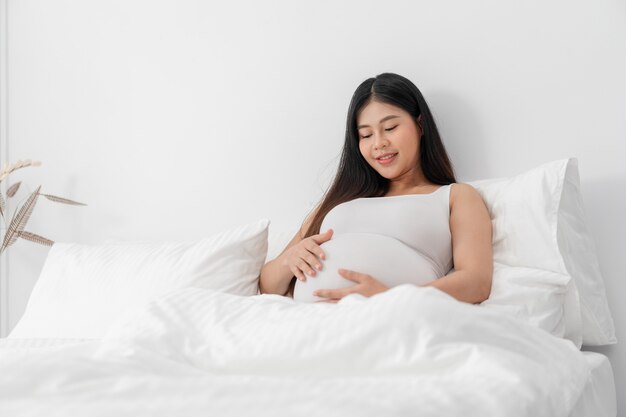 La donna incinta felice asiatica sta sedendosi sul letto e sta toccando la sua pancia