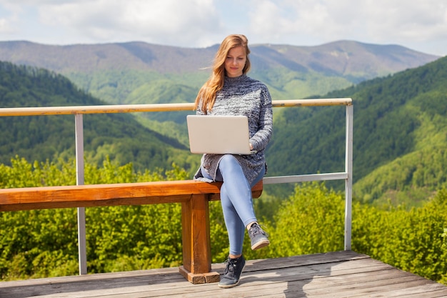 La donna incinta sta cercando qualcosa nel suo laptop che si siede su una panchina sullo sfondo della natura