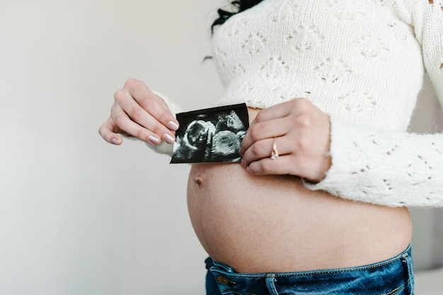 La donna incinta tiene la foto o l'immagine sulla pancia o l'ecografia sullo stomaco della futura figlia o figlio a casa Ragazza che aspetta il neonato Concetto di maternità Baby Shower