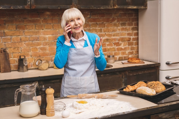 La donna invecchiata senior attraente sta cucinando sulla cucina. Nonna che produce una cottura saporita. Usando il telefono.