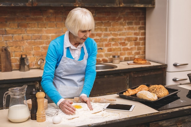 La donna invecchiata senior attraente sta cucinando sulla cucina. Nonna che produce una cottura saporita. Utilizzando un tablet.