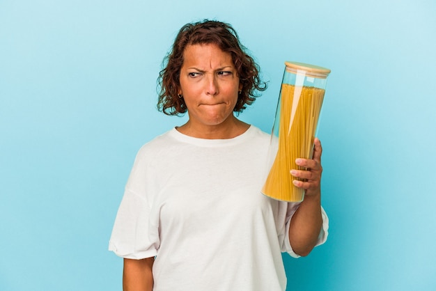 La donna latina di mezza età che tiene il barattolo di pasta isolato su sfondo blu confuso, si sente dubbiosa e incerta.