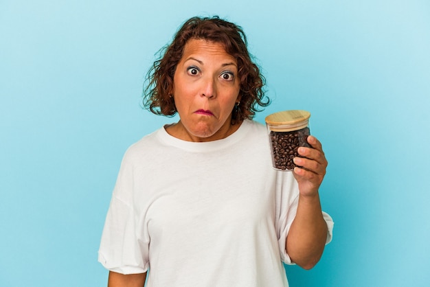 La donna latina di mezza età che tiene un barattolo di caffè isolato su sfondo blu alza le spalle e apre gli occhi confusi.