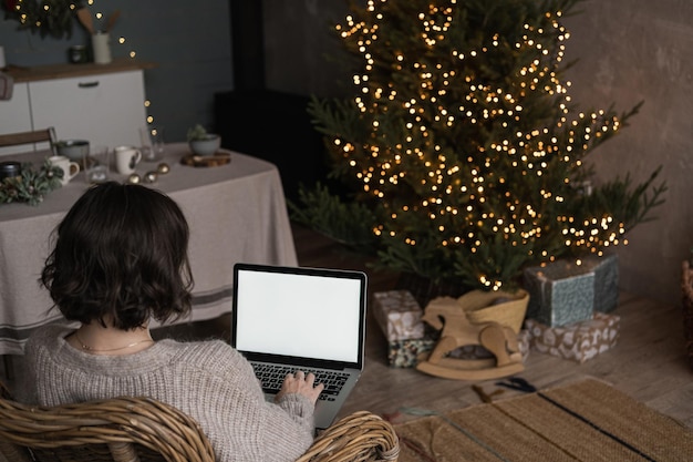 La donna lavora sul computer portatile con lo spazio vuoto della copia del mockup dello schermo Interno moderno del salone della casa decorato per la celebrazione di Natale con l'albero di Natale e le luci della ghirlanda
