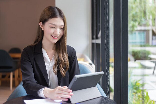 La donna lavoratrice professionista asiatica in abito nero sta lavorando su un tablet sul tavolo sorridendo felicemente