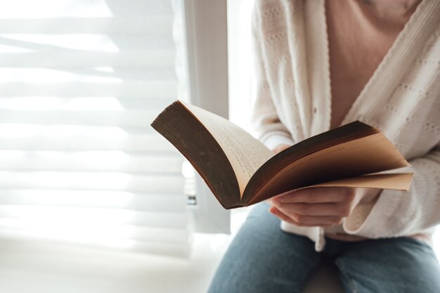 La donna legge un libro a casa vicino alla finestra durante il giorno.