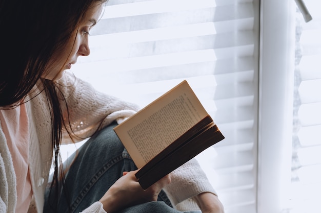 La donna legge un libro a casa vicino alla finestra durante il giorno.