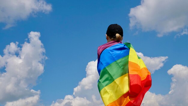 La donna lesbica bisessuale transgender sta con la bandiera LGBT contro il cielo blu con nuvole in una giornata di sole e celebra la giornata della bisessualità o la Giornata nazionale del coming out