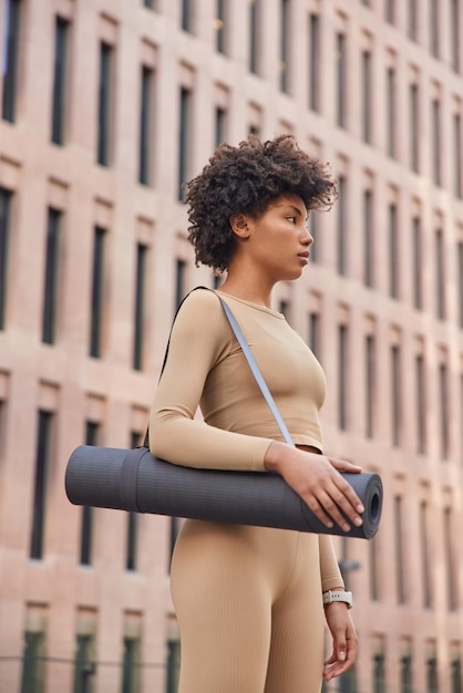 La donna magra dai capelli ricci si è concentrata seriamente sulla distanza vestita con una tuta da ginnastica beige portata in pose karemat arrotolate vicino all'edificio della città si prepara per l'allenamento all'aperto. Il modello femminile sportivo pratica lo yoga