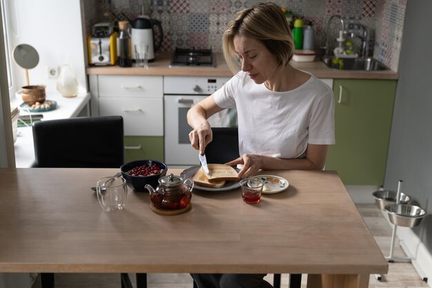 La donna matura prepara la colazione diffondendo toast con burro per bere il tè in una cucina accogliente