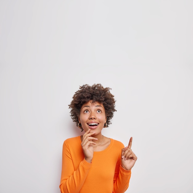 la donna mostra lo spazio della copia sopra ha gli occhi pieni di sorrisi di interesse indossa ampiamente un maglione arancione casual isolato su bianco. Guarda questo.