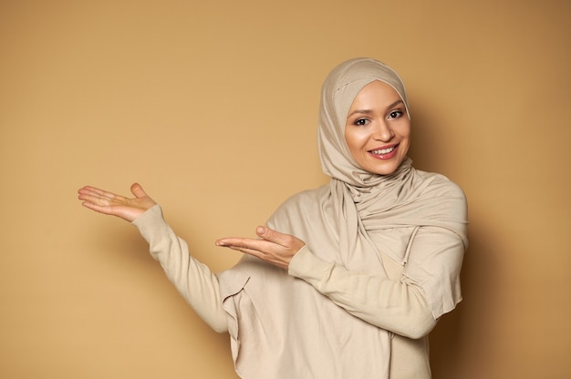 La donna musulmana con la testa coperta in hijab punta lateralmente sulla superficie beige e sorride con un sorriso a trentadue denti mentre guarda davanti