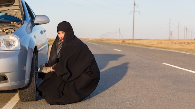 La donna musulmana in abito nazionale cambia la ruota di un'auto.