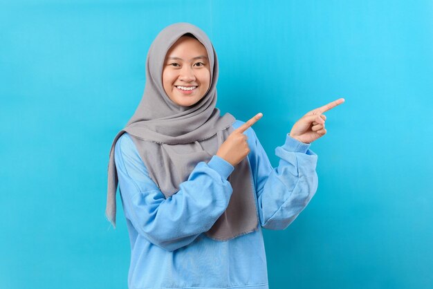 La donna musulmana indonesiana punta lontano sullo spazio della copia indossa un maglione blu ha un sorriso piacevole si sente ottimista isolato su sfondo blu