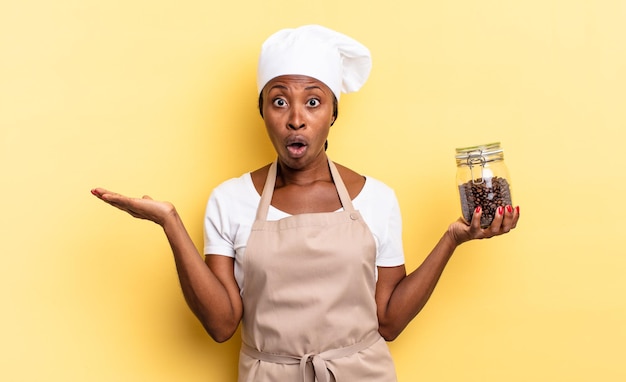 La donna nera dello chef afro che sembra sorpresa e scioccata con la mascella caduta tenendo un oggetto con una mano aperta sul concetto di chicchi di caffè laterali