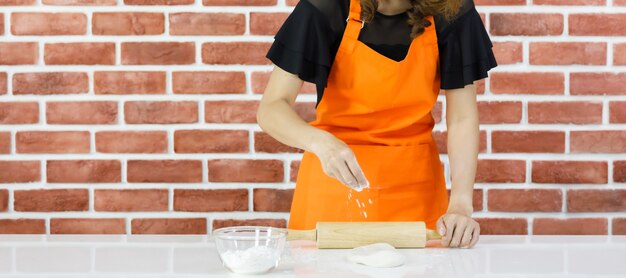 La donna non riconosciuta come chef in grembiule arancione si diverte a esercitarsi per impastare la pasta sul tavolo vicino al muro di mattoni con il mattarello di legno e una ciotola di vetro di farina in polvere all'interno della cucina di casa.