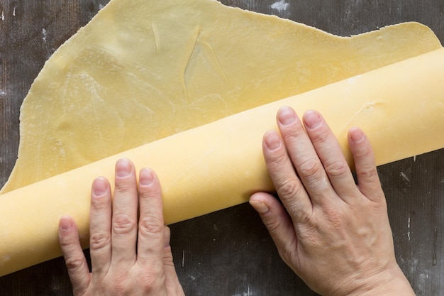 La donna passa la pasta per le tagliatelle con il mattarello sulla superficie di legno in polvere con farina