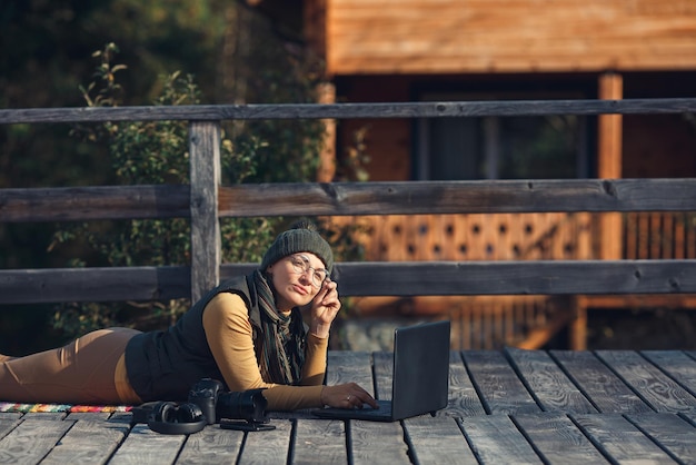 La donna pensierosa con gli occhiali si trova su una terrazza in legno e lavora su un laptop freelance concetto di lavoro a distanza su Internet