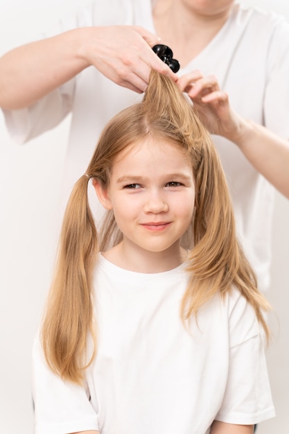 La donna pettina e fa i capelli di una bambina su uno sfondo bianco. La mamma è una parrucchiera. risparmiare denaro in un salone di bellezza. shampoo e cosmetici per i capelli dei bambini.