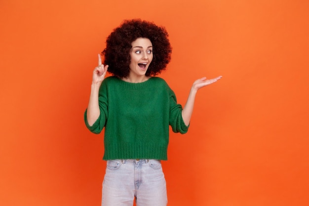 La donna positiva con l'acconciatura afro che indossa un maglione verde in stile casual che punta il dito verso l'alto ha un'idea che presenta un'area per la pubblicità Studio indoor girato isolato su sfondo arancione