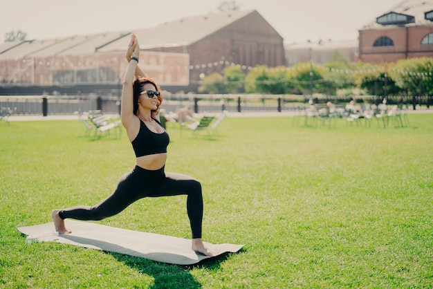 La donna positiva concentrata alza le braccia fa esercizi di pilates vestiti con occhiali da sole di abbigliamento attivo si allena durante le pratiche estive pose yoga sull'erba verde Concetto di persone e stile di vita sano