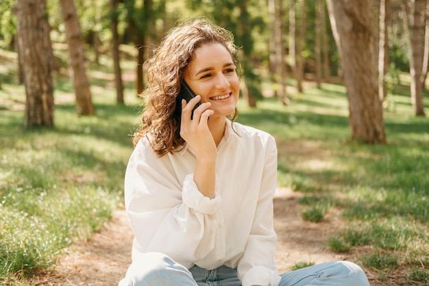 La donna riccia allegra sta avendo una telefonata mentre si gode seduta in una foresta