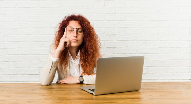 La donna riccia della giovane testarossa che lavora con il suo computer portatile che indica il tempio con il dito, pensando, si è concentrata su un compito.