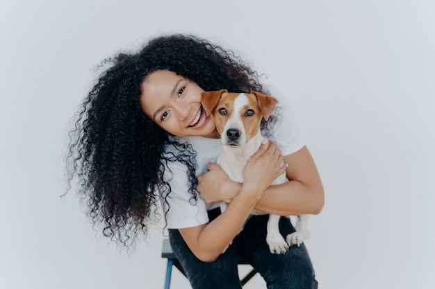 La donna riccia felice inclina la testa tiene il cane di razza ha un'espressione allegra sorride piacevolmente ha l'acconciatura riccia indossa una maglietta bianca isolata signora afro che accarezza l'animale domestico preferito