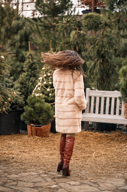 La donna riccia felice sorridente in una pelliccia sta girando tra gli alberi di Natale sulla strada in attesa di Natale e Capodanno