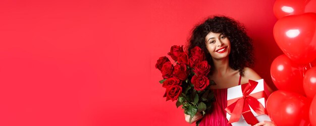 La donna romantica riceve bouquet di fiori e regali in vacanza in piedi vicino a palloncini a cuore e b rossa