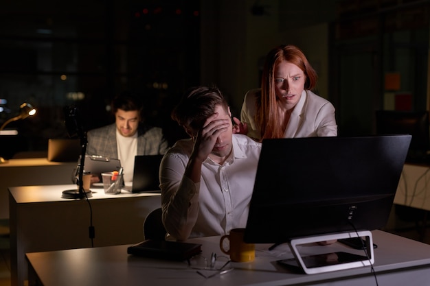 La donna rossa ansiosa preoccupata spaventata è insoddisfatta del lavoro di un uomo in ufficio, la donna è irritata guardando lo schermo del computer pc. vista laterale. a tarda notte in ufficio