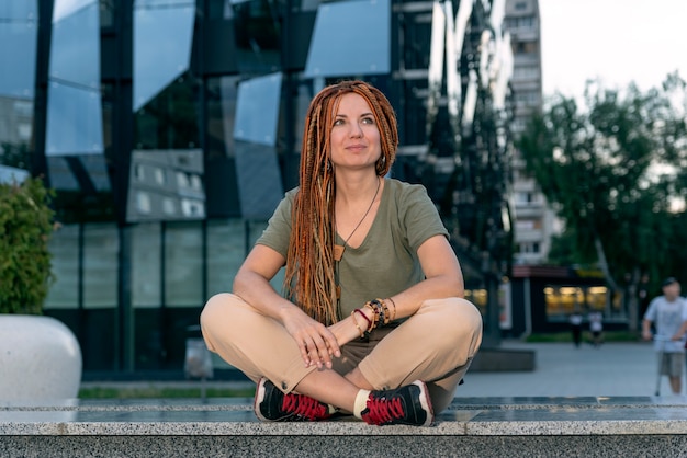 La donna rossa con lunghi dreadlocks siede nella posa del loto sullo sfondo di un edificio nero con finestre non standard.