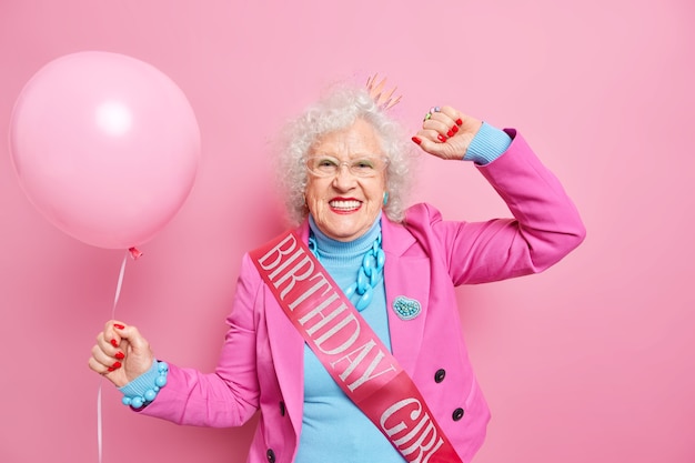 La donna rugosa dai capelli grigi spensierata balla sorrisi spensierati vestita positivamente in abiti festivi indossa il nastro di compleanno tiene il palloncino gonfiato