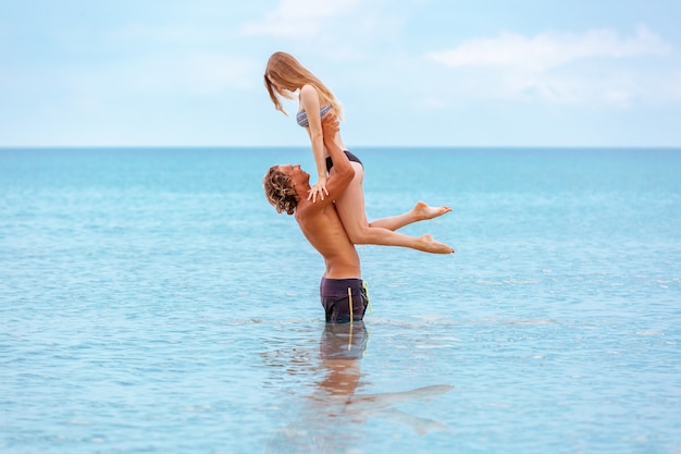 La donna salta all'uomo tra le sue braccia, in piedi nel mare. Entrambi sono in costume da bagno. Giovani coppie allegre sorridenti nell'amore divertendosi alla spiaggia sabbiosa.