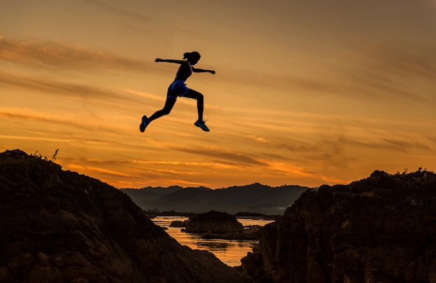 La donna salta attraverso lo spacco fra hill.woman che salta sopra la scogliera sulla priorità bassa di tramonto