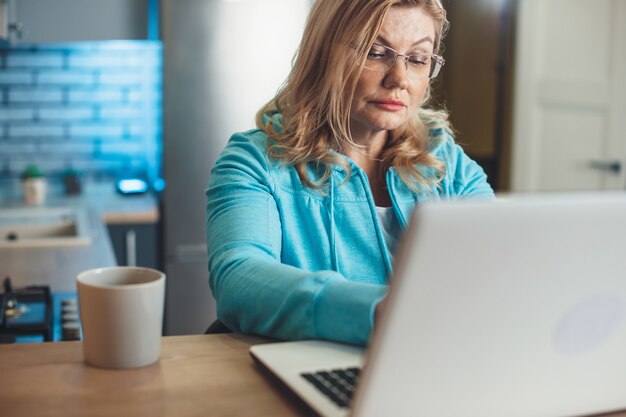 La donna senior bionda sta lavorando al laptop da casa bevendo una tazza di tè in cucina