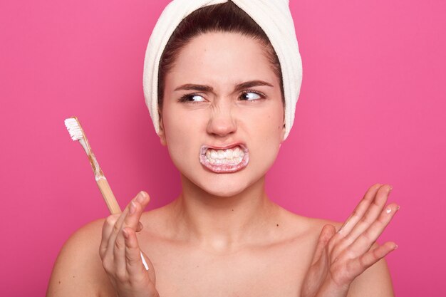 La donna seria attraente con capelli scuri e l'asciugamano bianco sulla testa, tenendo il toothbrushe in mano, ha espressione facciale arrabbiata, guardando da parte