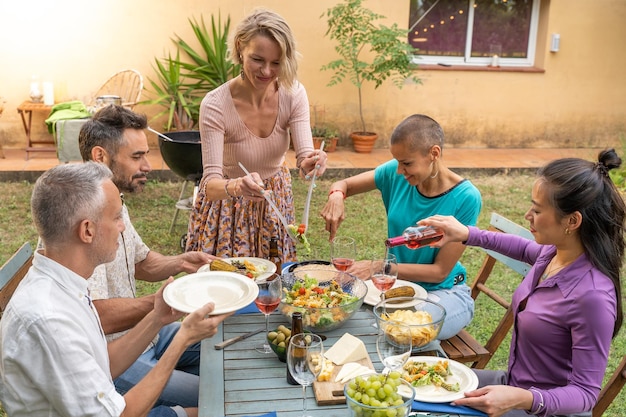 La donna serve il cibo nel patio della casa Amici felici che ridono intorno al tavolo all'aperto