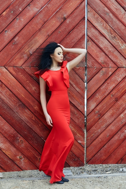 La donna sexy del brunette in un vestito rosso sta levandosi in piedi vicino al cancello rosso