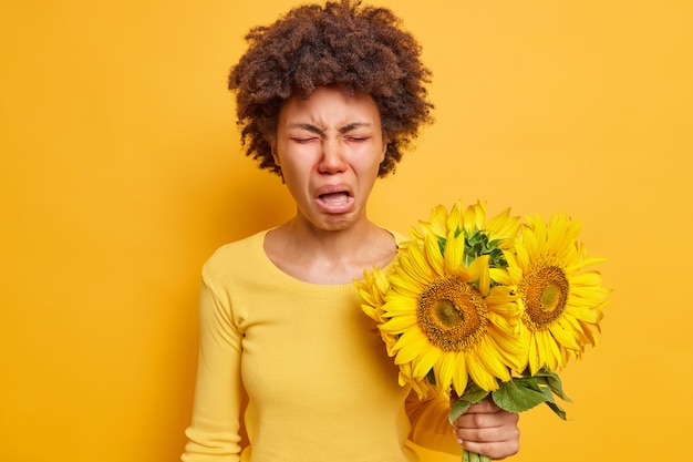 la donna si imbroncia e si acciglia il viso ha una smorfia infelice tiene un mazzo di girasoli che causano una reazione allergica indossa un maglione casual isolato su un giallo vivido