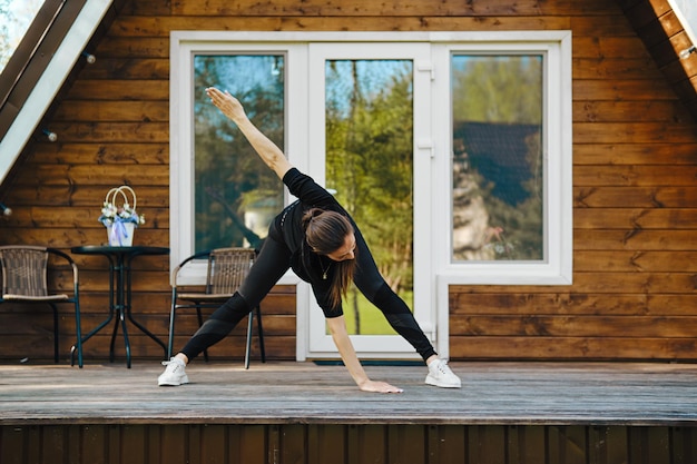 la donna si piega e allarga le braccia ai lati durante l'allenamento mattutino sulla terrazza della casa di legno