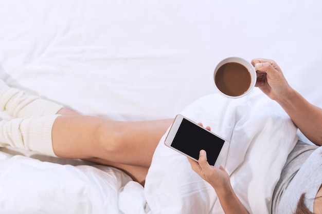 La donna si rilassa sul letto mentre usa il telefono cellulare e una tazza di caffè in mano, vista dall'alto e copia spazio.