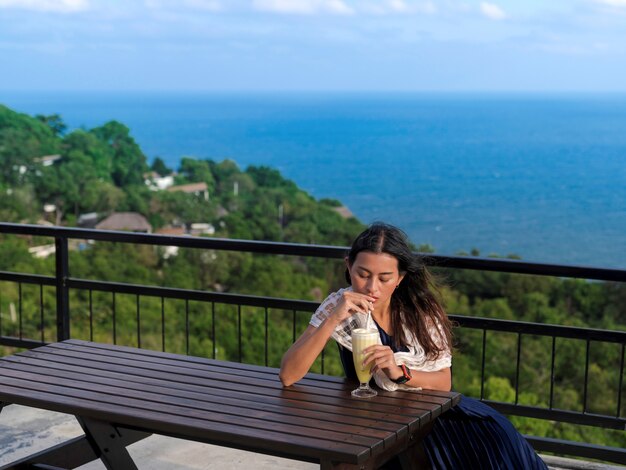 La donna si siede al tavolo di legno bevendo frappe di ananas sulla terrazza con vista mare sul picco di montagna a Koh Tao island, Suratthani, Thailandia