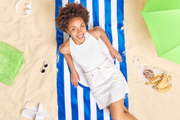 la donna si siede su un asciugamano a righe sembra felicemente vestita in abito estivo trascorre le vacanze estive in spiaggia prende il sole e si sente rilassata. Il miglior concetto di vacanza.