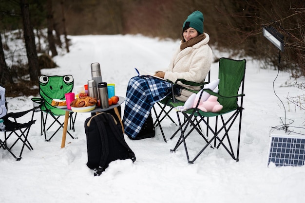 La donna si siede sulla sedia e legge il libro nella foresta invernale durante un picnic