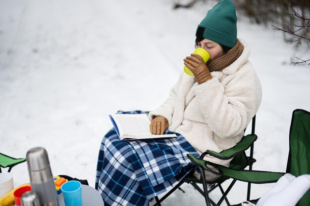 La donna si siede sulla sedia, legge il libro e beve il tè nella foresta invernale durante un picnic