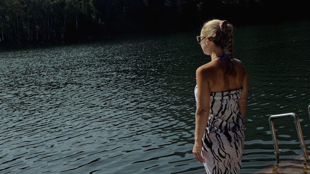 La donna si trova su un lettino con occhiali da sole e uno scialle di seta boho La ragazza riposa su un molo sottomarino di legno inondato Il marciapiede è coperto d'acqua nel lago Sullo sfondo ci sono una montagna e una foresta