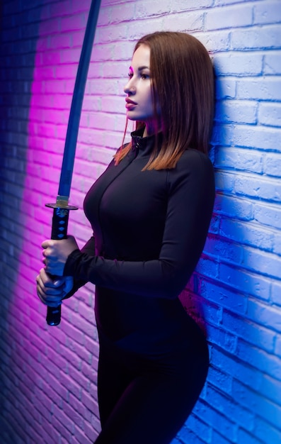 La donna snella sexy in abiti neri con una katana in mano alla luce al neon