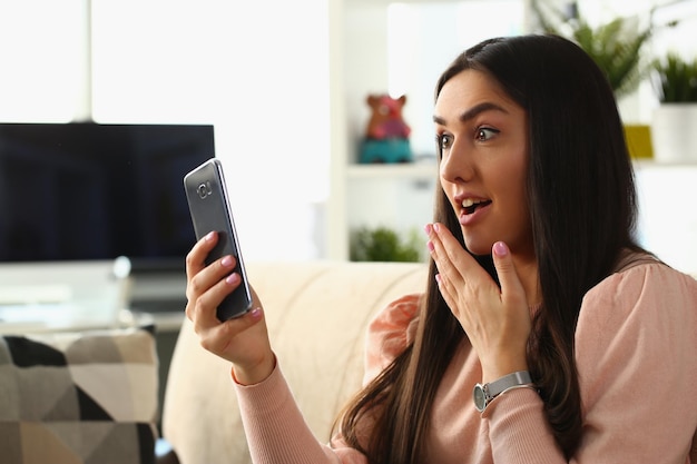 La donna sorpresa dallo shock guarda lo schermo dello smartphone mentre è seduta sul divano di casa